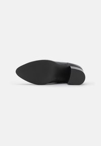 Zapato de vestir negro y elegante con punta vista desde el lado de la suela, mostrando la suela texturizada y el tacón bajo en un fondo blanco.