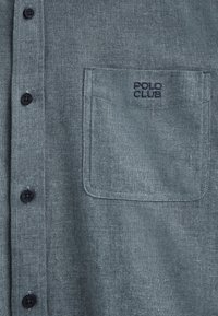 Grå skjorta med knappar i svart, med en bröstficka broderad med "POLO CLUB." Tyget har en texturerad yta.