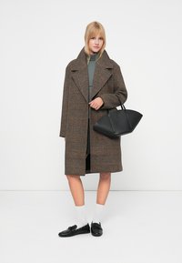 Bruin geruit overjas met brede revers, zwarte handtas, grijze turtleneck en zwarte instappers. Model draagt witte sokken en staat tegen een witte achtergrond.