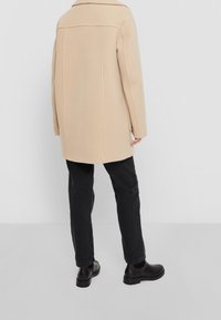 Abrigo de lana beige con corte recto, costuras traseras y cuello con solapa, combinado con pantalones negros y botines negros.