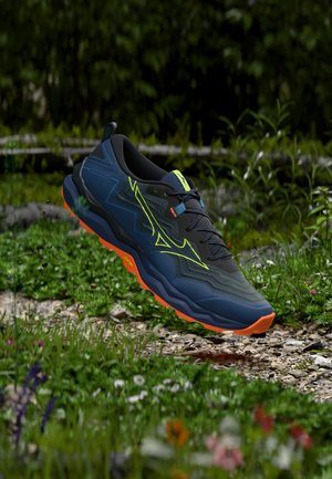 Zapatilla de trail azul oscuro con detalles en amarillo neón y suela naranja, flotando sobre un camino de grava rodeado de hierba verde y flores silvestres.