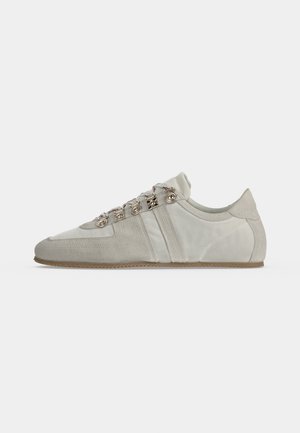 Zapatilla deportiva de perfil bajo en gris claro con detalles de ante, ojales plateados y suela fina de goma, mostrada desde el lado externo sobre fondo blanco.