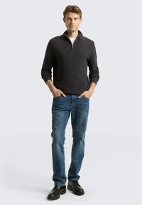 Dunkelgrauer Pullover mit Reißverschluss und strukturiertem Strick, kombiniert mit blauen Jeans und schwarzen Schuhen. Mann steht mit den Händen in den Taschen vor einem hellen Hintergrund.