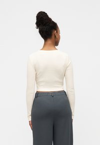 Ελαφρώς μπεζ κοντό τοπ με μακριά μανίκια και στρογγυλή λαιμόκοψη, σε συνδυασμό με ψηλόμεσα σκούρα γκρι παντελόνια. Λείο ύφασμα με ελάχιστες λεπτομέρειες.