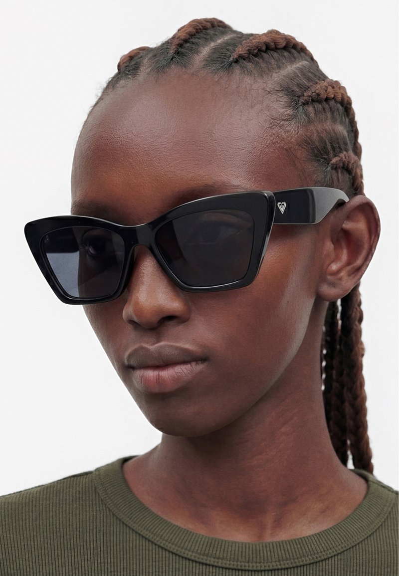 Personne aux cheveux étroitement tressés portant des lunettes de soleil noires de style œil de chat et un t-shirt côtelé vert olive sur fond blanc.
