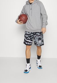 Felpa grigia con cappuccio e coulisse, pantaloni corti neri e grigi con motivo e logo "Raiders", scarpe da ginnastica bianche con accenti blu.