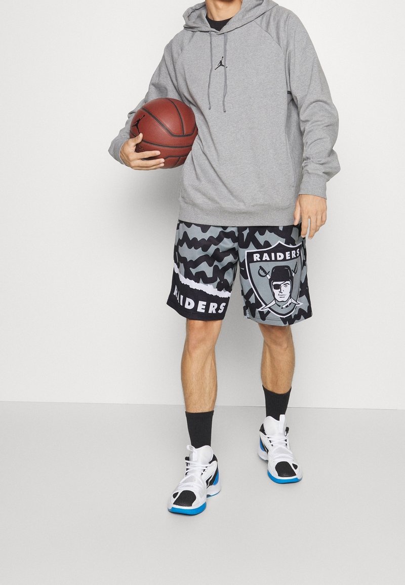 Felpa grigia con cappuccio e coulisse, pantaloni corti neri e grigi con motivo e logo "Raiders", scarpe da ginnastica bianche con accenti blu.