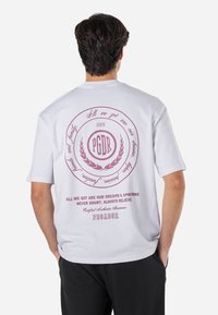Camiseta blanca de manga corta con un gráfico circular en rosa oscuro que incluye texto, acentos de laurel y un ajuste holgado.