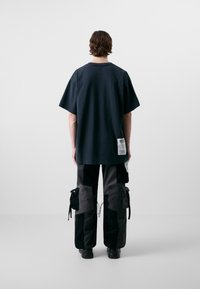 Camiseta negra de gran tamaño con mangas cortas y una etiqueta en el lado inferior, combinada con pantalones cargo holgados de varios bolsillos en tonos oscuros.