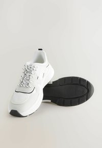 Baskets de sport blanches avec une tige en mesh texturé, accents noirs et semelle en caoutchouc à motifs. Comprend des lacets et une languette pour un enfilage facile.
