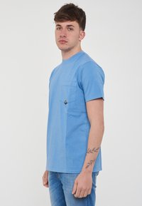 T-shirt di cotone azzurro chiaro con collo rotondo, maniche corte e taschino sul petto con etichetta nera triangolare. Texture liscia.