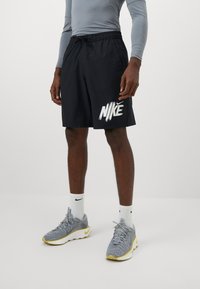 Vyras vilkintis juodus Nike šortus, pilkus sportinius batus su geltonais padais, baltas Nike kojines ir pilką ilgomis rankovėmis marškinį, stovintis ant balto grindų.