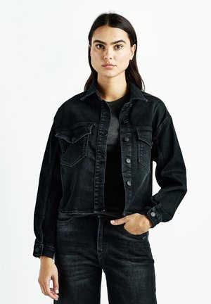 Young woman with long dark hair wearing a black denim jacket and black jeans, standing against a plain white background.