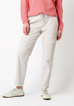 Osoba ubrana w jasnobeżowe spodnie typu cargo ze ściągaczem w pasie, białe casualowe buty i koralową bluzkę z długim rękawem, stojąca na białym tle.