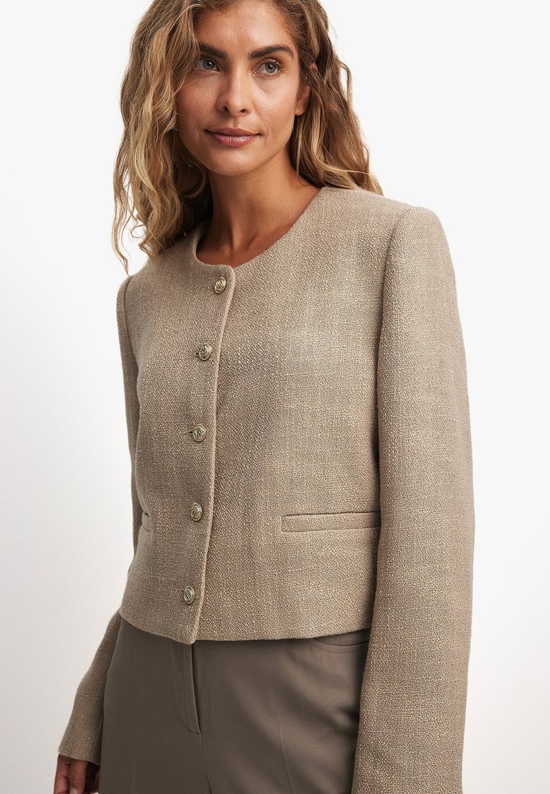 Bežs teksturēts apgriezts žakets ar apaļu kakla izgriezumu, piecu pogu aizdare un priekšējām kabatām, apvienots ar brūniem biksēm. Piezīme: auduma vieglais mirdzums.