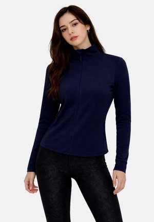 Junge Frau mit langen braunen Haaren, die eine marineblaue Reißverschlussjacke und schwarze Hose trägt, steht vor einem weißen Hintergrund.
