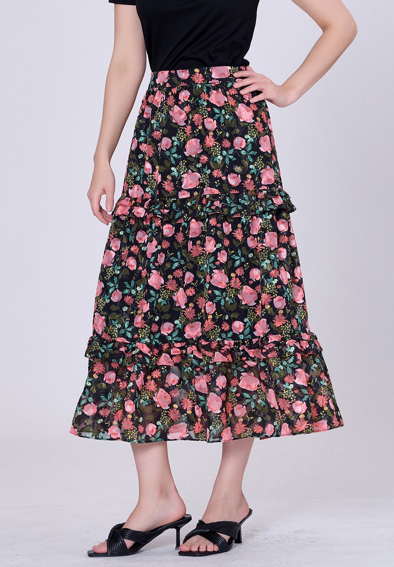 Jupe maxi florale avec fond noir, volants et motifs de fleurs roses et vertes. Associée à des sandales noires à talons.