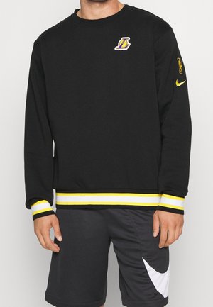 Man draagt een zwart Los Angeles Lakers-sweatshirt met lange mouwen, gele-witte gestreepte manchetten en zwarte Nike-shorts met een grote witte swoosh.