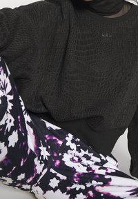 Černá mikina s hadí texturou, zdobená vyšitým logem, doplněná legínami s květinovým potiskem v černé, bílé a fialové barvě.