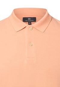 Pfirsichfarbiges Poloshirt aus Baumwolle, klassischer Kragen, zwei Knöpfe an der Knopfleiste, strukturiertes Gewebe mit subtiler Rippung, Etikett innen am Kragen sichtbar.