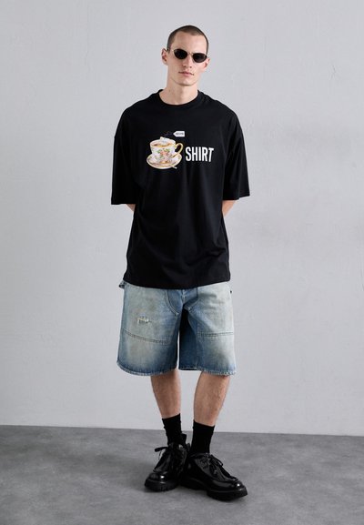 T-shirt noire surdimensionnée avec un graphique de tasse et le texte "SHIRT" ; shorts en denim décolorés ; chaussures noires ; accessoires minimalistes, fond uni.