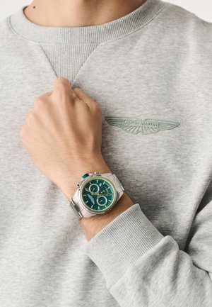 Zilveren chronograafhorloge met een blauwgroene wijzerplaat, gele accenten en drie subwijzerplaten, gedragen aan een pols naast een grijze trui.