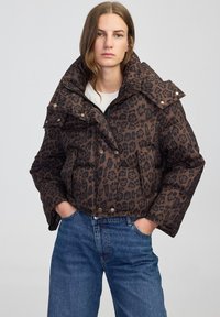 Bruine pufferjack met luipaardprint, voorzien van een hoge kraag, drukknoopsluiting en zijzakken, gecombineerd met blauwe jeans.