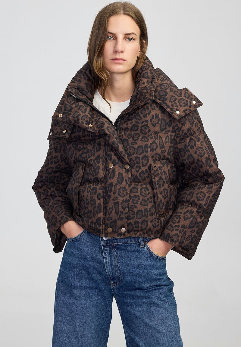 Bruine pufferjack met luipaardprint, voorzien van een hoge kraag, drukknoopsluiting en zijzakken, gecombineerd met blauwe jeans.