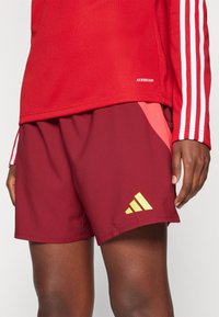 Červený športový top s bielymi pruhmi a štítkom „AEROREADY“. Bordové šortky s žltým logom Adidas a bočnými panelmi z priedušnej sieťoviny.
