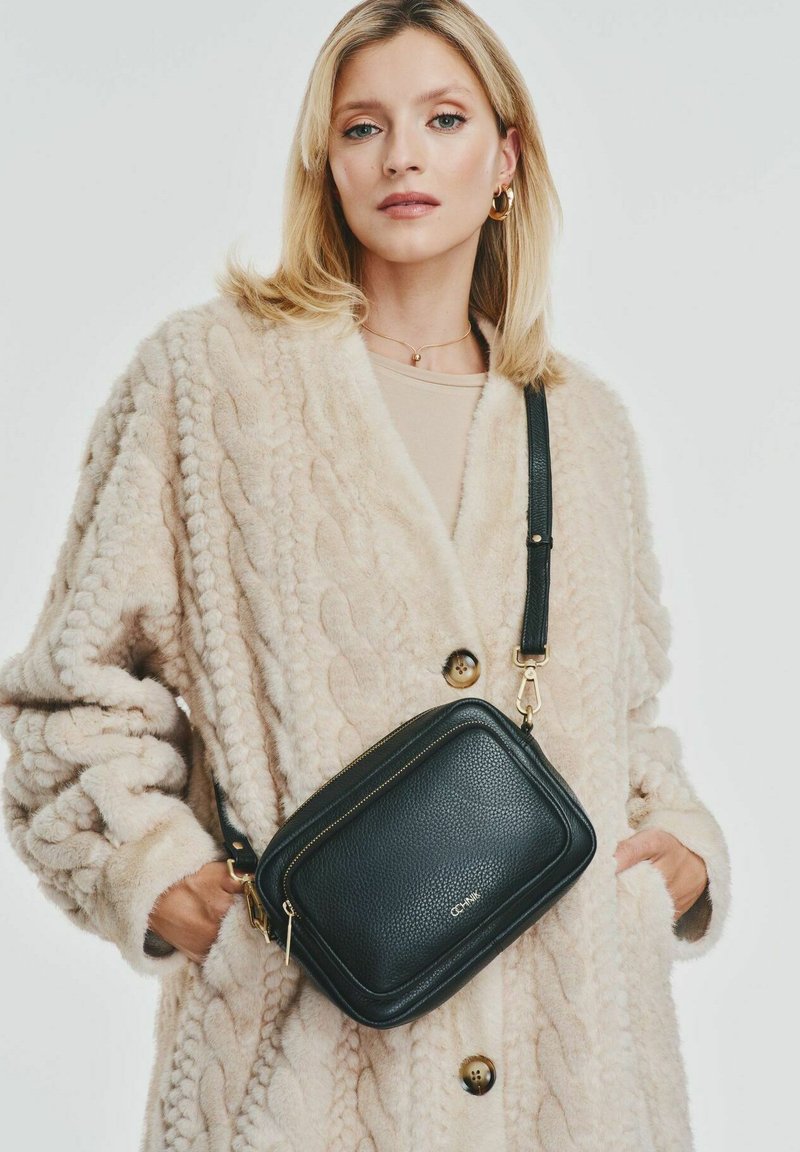 Czarna skórzana torebka crossbody o teksturowanej powierzchni, z zapięciem na zamek błyskawiczny i złotymi elementami, noszona na kremowej kardiganowej bluzie w warkocze.