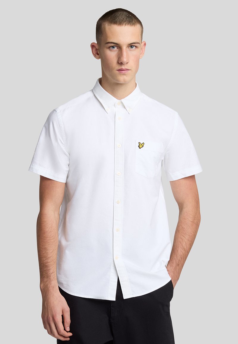 Camicia bianca a maniche corte in cotone, con colletto a punta, taschino sul petto e una piccola etichetta gialla sul taschino.