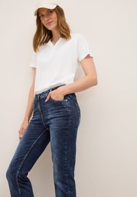 Witte short-sleeve v-hals t-shirt gecombineerd met donkerblauwe high-waisted jeans, met een zak en lichte vervaging, gedragen met een beige baseballcap.