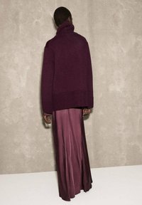 Maglione a collo alto oversize di un profondo bordeaux con polsini a costine, abbinato a una lunga gonna in satin bordeaux fluente. Sfondo opaco.