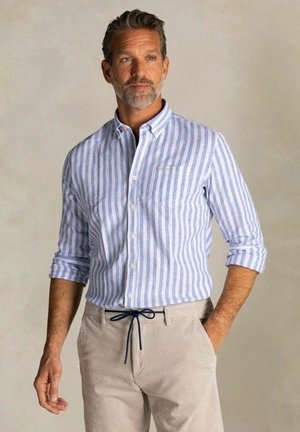 Man met grijze baard, gekleed in een blauw-wit gestreept overhemd met knopen en beige broek met een donkere trekkoord.