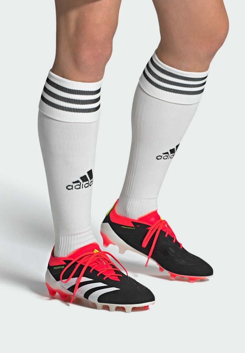 Scarpe da calcio nere e rosse con strisce bianche, dotate di una tomaia texturizzata, stringhe rosse brillanti e calze bianche con strisce nere.