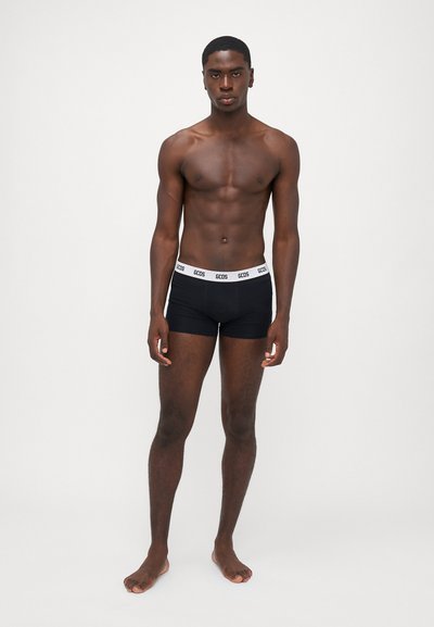 Schwarze Baumwollboxershorts mit einem weißen Bund, auf dem "GCDS" steht. Das Model hat einen durchtrainierten Körper und steht barfuß vor einem schlichten Hintergrund.