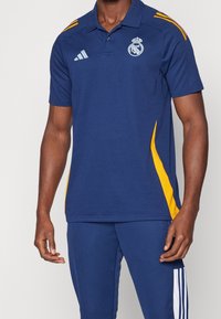 Homme portant un polo Real Madrid bleu et jaune et un pantalon Adidas bleu avec des rayures blanches sur le côté, debout devant un fond uni.