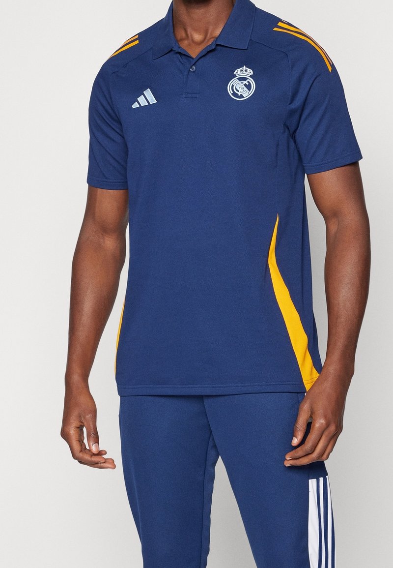 Homme portant un polo Real Madrid bleu et jaune et un pantalon Adidas bleu avec des rayures blanches sur le côté, debout devant un fond uni.