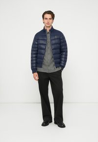 Tengerészkék steppelt dzseki függőleges párnázott részekkel, magas gallérral, ezüst cipzárral, szürke gombos ing és fekete nadrág fölött viselve.