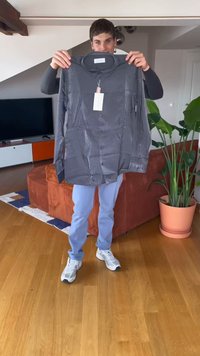 Person steht drinnen und hält in einem Wohnzimmer eine dunkelgraue Langarmhemd mit Etiketten hoch.