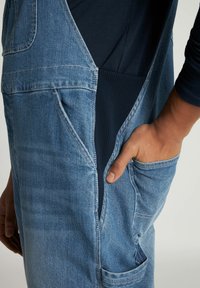 Salopette in denim blu con una finitura testurizzata, dotata di tasche laterali e una sezione a coste blu navy per il comfort. Spalline regolabili visibili.