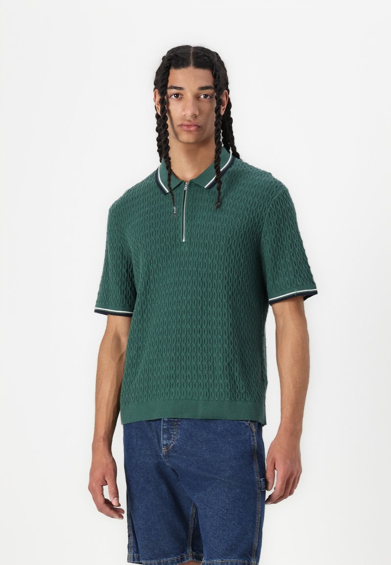 Polo verde a maniche corte con un motivo testurizzato, colletto a coste con dettagli bianchi e blu marino, abbinato a pantaloni corti di denim.