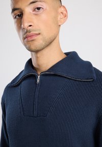 Young man wearing a dark blue knitted sweater with a half-zip collar, looking slightly away, against a plain light background.