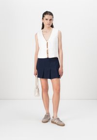 Witte, mouwloze crop top met een diepe V-hals en strikdetail, gecombineerd met een marineblauwe geplooide rok en beige sneakers. Kleine lichte tas erbij.