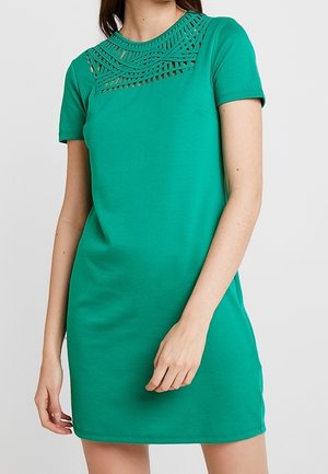 Frau trägt ein grünes, kurzärmeliges Kleid mit einem geometrischen Cut-out-Muster über dem oberen Brustbereich und dem Ausschnitt.