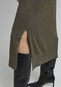 Robe tricotée verte olive avec une fente sur le côté, associée à des bottes noires en cuir montantes, mettant en valeur la texture et le contraste entre les matériaux.