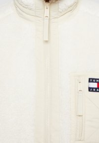 Kremowa kurtka z polaru z wysokim kołnierzem, przednim zamkiem błyskawicznym i boczną kieszenią. Posiada małą naszywkę z logo w czerwonych, białych i niebieskich akcentach.