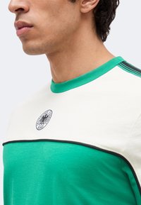 Κοντινό ενός ατόμου που φοράει πράσινο και άσπρο αθλητικό μπλουζάκι με μαύρο λογότυπο Deutscher Fußball-Bund στο στήθος.