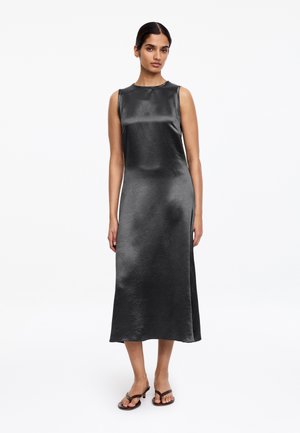 Vrouw draagt een mouwloze, enkellange zwarte satijnen jurk met een ronde halslijn en zwarte sandalen met hakken, staande tegen een witte achtergrond.