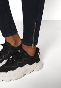 Svarta sneakers med vita sulor, bärs med mörk denimjeans med en silverdragkedja vid vristen för justerbar passform. Slät textur.